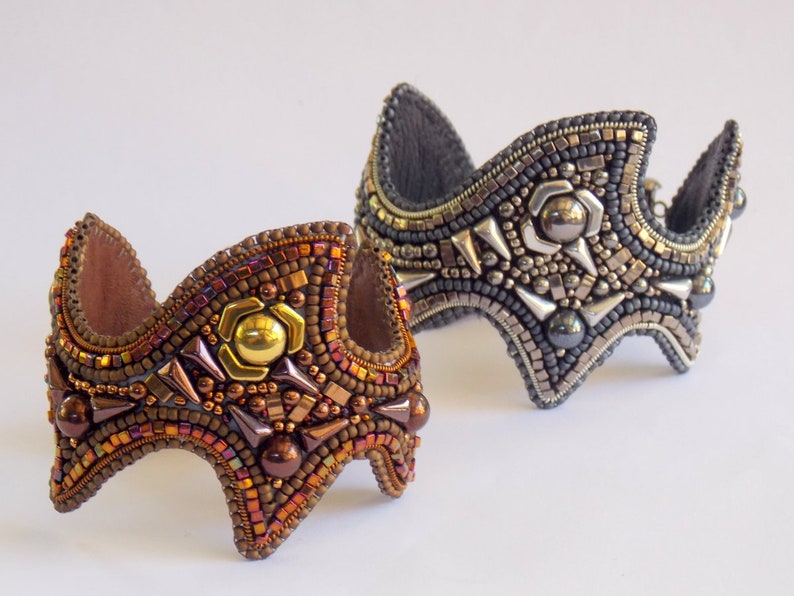 May include: Two beaded cuff bracelets. One is bronze and gold, with a central gold accent. The other is silver and grey, with silver spikes and a central silver accent. Both have a curved shape and a leather backing.