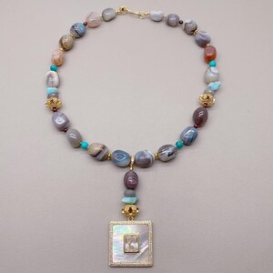 May include: A beaded necklace featuring a variety of natural stone beads in shades of gray, brown, and tan, interspersed with turquoise beads and gold-colored accents. The pendant is a square shape with a mother-of-pearl center and a clear stone.