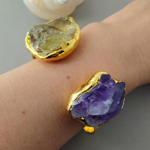 May include: Two gold-plated bracelets, each featuring a large, raw gemstone. One bracelet has a light green stone, and the other has a deep purple amethyst. The bracelets are set against a neutral background.