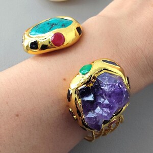 May include: Gold-toned jewelry with gemstone accents. One piece features a turquoise stone with ruby and black accents. The other has a large purple amethyst stone with green and black accents. Both are on a wrist.