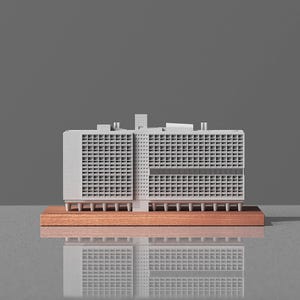 May include: A miniature architectural model of a building with a grid-like facade. The model is light gray and sits on a rectangular, reddish-brown wooden base. The building has multiple levels and a flat roof. The background is a solid gray.