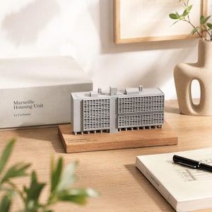 Könnte beinhalten: Graues Architekturmodell der Unité d'Habitation, auch bekannt als Marseille Housing Unit, von Le Corbusier. Das Modell steht auf einem Holzsockel und wird von einer Box und einem Buch begleitet. Das Modell ist etwa 15 cm lang.