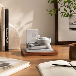 Puede incluir: Una maqueta arquitectónica del Museo Guggenheim de Nueva York. El modelo es de color gris claro y se asienta sobre una base de madera oscura. Libros y una foto enmarcada se encuentran en el fondo.