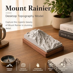 May include: A desktop topography model of Mount Rainier, a detailed white mountain replica on a wooden base. The model is displayed on a desk with a compass, books, and a plant. Text on the image reads "Mount Rainier Desktop Topography Model".