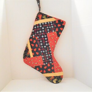 May include: A red, black, and gold Christmas stocking with a patchwork design featuring snowflakes, Christmas trees, and the words "Merry Christmas".