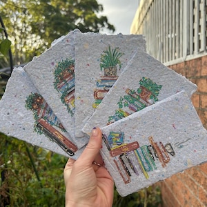 May include: A collection of handmade paper cards with illustrations of books and plants. The cards are light gray with colorful flecks and feature watercolor-style drawings of stacked books, potted plants, and greenery. The cards are held in a person's hand.