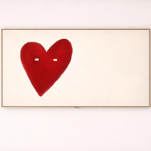 May include: A framed artwork featuring a large, red heart with two small white eye-like shapes. The heart is centered on a cream-colored background, encased in a thin, gold-toned frame. The artwork is mounted on a white wall.