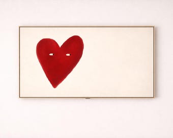 May include: A framed artwork featuring a large, red heart with two small white eye-like shapes. The heart is centered on a cream-colored background, encased in a thin, gold-toned frame. The artwork is mounted on a white wall.