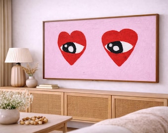 Könnte beinhalten: Ein gerahmtes Kunstwerk mit zwei roten herzförmigen Augen auf rosa Hintergrund. Das Kunstwerk wird über einem Holzschrank mit gewebten Paneelen ausgestellt. Eine Lampe und eine Vase stehen auf dem Schrank, und ein Couchtisch befindet sich davor.