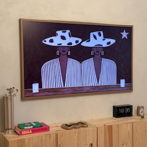 May include: A framed art print featuring two figures wearing white hats with black spots and striped tops. The artwork is displayed above a wooden cabinet with books and decorative items.