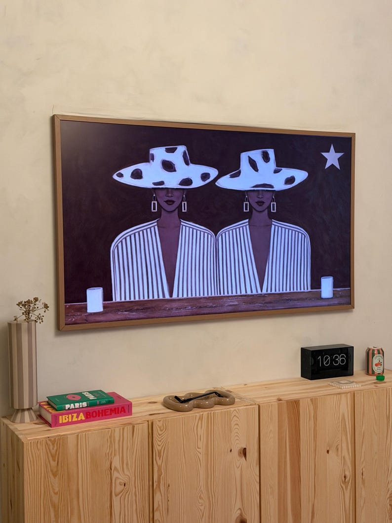 May include: A framed art print featuring two figures wearing white hats with black spots and striped tops. The artwork is displayed above a wooden cabinet with books and decorative items.