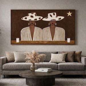 May include: A framed artwork depicts two figures wearing hats with a cowhide pattern. They are dressed in striped jackets, with a star and two candles in the background. The artwork is displayed above a gray sofa.
