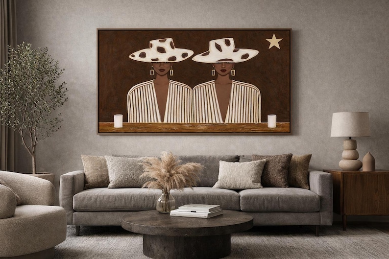 May include: A framed artwork depicts two figures wearing hats with a cowhide pattern. They are dressed in striped jackets, with a star and two candles in the background. The artwork is displayed above a gray sofa.
