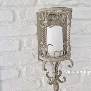 May include: A distressed, off-white metal wall sconce with a cylindrical candle holder. The sconce features ornate scrollwork and a decorative base. A white pillar candle sits inside the holder. The background is a white brick wall.