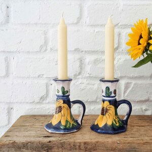 May include: Two ceramic candle holders with handles, each featuring a hand-painted yellow sunflower design on a dark blue base. Each holder contains a tall, slender, cream-colored taper candle. The holders are positioned side by side.