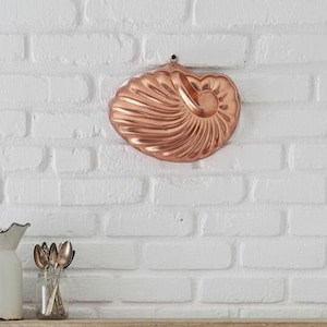 May include: A copper-colored, nautilus-shaped mold hangs on a white brick wall. Below, a wooden shelf holds a white pitcher, a small glass jar with spoons, and a lower shelf with hooks. The kitchen decor has a rustic, vintage aesthetic.