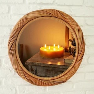 May include: A round wall mirror featuring a thick, braided natural rattan frame. The reflective surface shows a reflection of a cylindrical yellow candle with four lit wicks resting on a wooden surface, along with a small potted plant and a book.