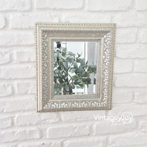 Op de afbeelding: Een vierkante, sierlijke spiegel met een crèmekleurige lijst hangt aan een witte bakstenen muur. De spiegel reflecteert een groene plant. De tekst "VintageyJoy" staat op de muur onder de spiegel.