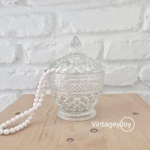 May include: A clear, vintage-style glass candy dish with a lid, featuring a diamond pattern. A white beaded necklace is draped over the dish. The dish is on a wooden surface against a white brick wall.