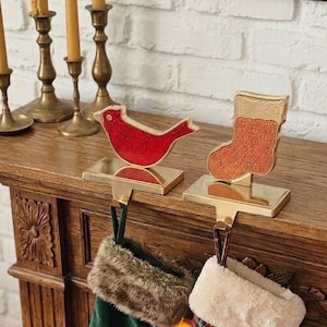Puede incluir: Dos soportes de metal dorado para calcetines navideños con formas decorativas. Uno muestra un pájaro cardenal rojo con un sutil patrón en relieve, mientras que el otro presenta una forma de calcetín naranja texturizado sobre bases rectangulares.