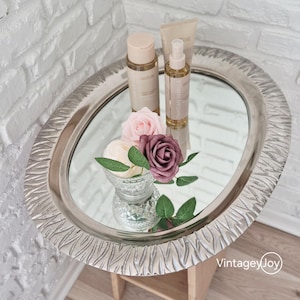 May include: An oval silver mirrored tray with a decorative edge holds beauty products and floral arrangements. The tray features a clear glass bowl with white and purple roses, and several bottles of skincare products. The tray sits on a small wooden table.
