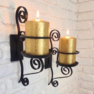 Peut inclure: Un bougeoir mural en métal noir avec deux bras ornés de volutes. Chaque bras supporte une bougie cylindrique dorée avec une flamme allumée. La structure est fixée sur une plaque rectangulaire noire contre le mur.