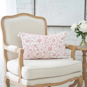 May include: A decorative rectangular pillow with a pink floral pattern rests on a cream-colored chair. The chair has a light wood frame and is next to a small wooden side table with a vase of white flowers.