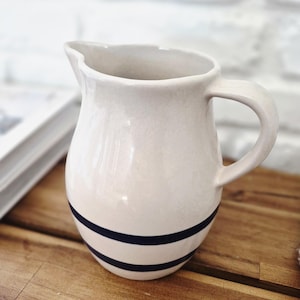 Puede incluir: Jarra de cerámica color crema con diseño de rayas azul oscuro. La jarra tiene un asa curva y un pico vertedor. Una pieza decorativa para la cocina o el comedor.