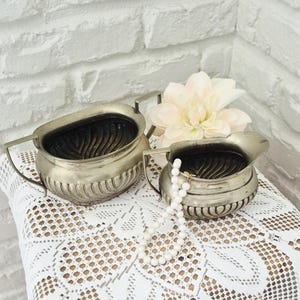 May include: A silver-toned metal sugar bowl and creamer set featuring a ribbed base design and two side handles. The interior of each vessel displays a textured, wave-like pattern. A string of white beads rests inside the creamer, and a pale flower sits behind.