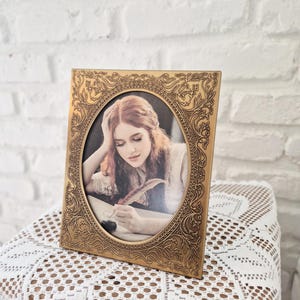 May include: A gold-colored, ornate picture frame with an oval opening. The frame features a detailed floral design around the edges. Inside the frame is a sepia-toned photograph of a person writing with a quill pen. The frame is displayed on a white lace tablecloth.
