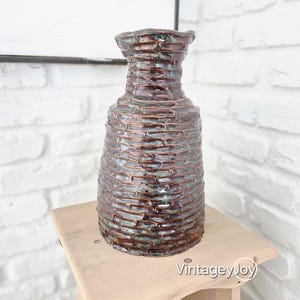 Puede incluir: Un jarrón de cerámica marrón texturizado con un diseño único y en capas. El jarrón tiene una base ancha y un cuello más estrecho, con un borde ligeramente irregular. La superficie tiene un brillo metálico y se exhibe sobre un soporte de madera.