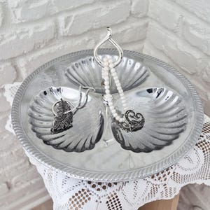 May include: A silver-toned metal serving tray featuring three shell-shaped compartments and a central handle. A white beaded necklace with a heart-shaped pendant and a silver-toned chain necklace with a leaf-shaped pendant rest inside the tray.