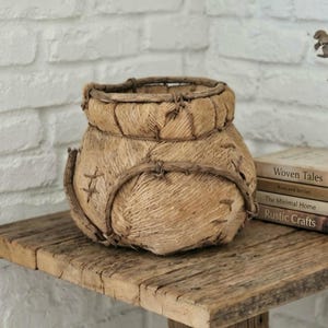 May include: A round, tan, woven jute basket featuring a thick, reinforced rim and decorative stitched detailing across its exterior surface. The basket has a textured, fibrous appearance with visible rope-like construction and a wide, open top.