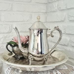 May include: A polished silver-plated teapot with an ornate handle and spout, featuring a matching lid with a finial. It sits on a round silver tray alongside a smaller silver sugar bowl containing a pink artificial rose with green leaves.