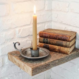 Puede incluir: Un candelabro de metal con asa curva y una vela cónica encendida, colocado junto a una pila de tres libros antiguos de tapa dura con lomos marrones desgastados, sobre un estante de madera rústica contra una pared de ladrillo blanco.
