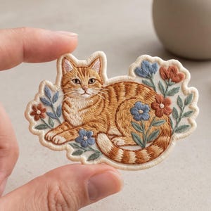 May include: An embroidered patch featuring an orange tabby cat lying down surrounded by small blue and rust-colored flowers with green leaves. The patch has a cream-colored fabric base with a scalloped edge and detailed thread work.