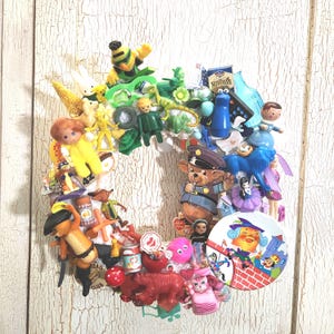 Pode incluir: Uma coroa colorida feita de vários brinquedos e figuras pequenas. A coroa apresenta uma variedade de cores, incluindo amarelo, verde, azul e vermelho. Os brinquedos incluem um urso com uniforme de polícia, um leão azul e uma boneca.