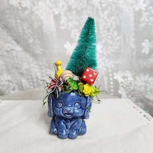 May include: A blue ceramic dog figurine serving as a planter, filled with a green bottle brush tree, a red die, a yellow game piece, a small green frog, a wooden bead, a red, white, and blue striped ribbon, and yellow fabric flower petals.