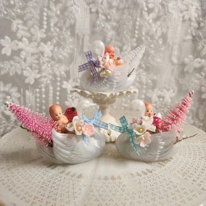 以下が含まれることがあります： 小さな人形と装飾要素が入った、白鳥の形をした3つの白い置物。白鳥はピンクのボトルブラシの木、花、パステルカラーのリボンで飾られています。1羽の白鳥が白い台座の上に置かれています。