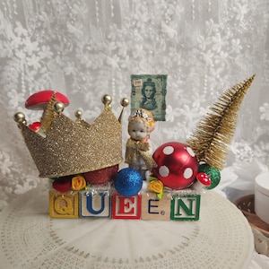 May include: A decorative arrangement featuring five wooden alphabet blocks spelling QUEEN, topped with a gold glitter crown, a small porcelain doll, a gold bottle brush tree, red and white polka dot ornaments, and a small green postage stamp.