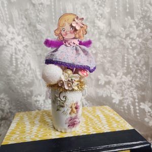 May include: A decorative arrangement in a vintage teacup, featuring a doll figure, glittery spheres, and floral accents. The doll has a floral dress and a flower in her hair. The teacup is decorated with pink and yellow roses.