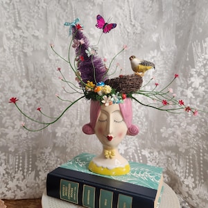 Puede incluir: Un jarrón de cerámica caprichoso con cabello rosado, ojos cerrados y labios rojos. El jarrón está adornado con un pájaro, un nido, una mariposa y detalles florales. Se encuentra sobre un libro vintage con una cubierta turquesa.