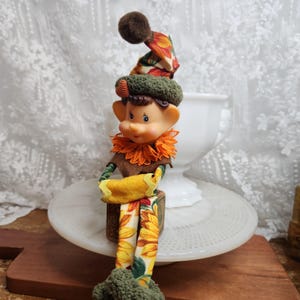 Könnte beinhalten: Eine kleine Elfenfigur mit einer spitzen Mütze, die einen braunen Bommel und ein Sonnenblumenmuster aufweist. Die Elfe hat ein pfirsichfarbenes Gesicht, spitze Ohren, einen orangefarbenen Spitzenkragen und Gliedmaßen mit Sonnenblumenmuster.