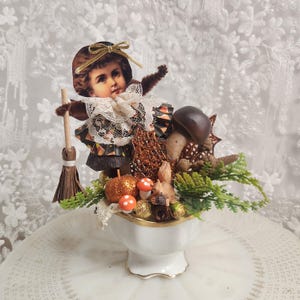 Puede incluir: Un arreglo caprichoso con temática de Halloween en un cuenco de porcelana blanca con ribete dorado. Presenta una imagen de estilo vintage de un niño disfrazado de bruja, una escoba, setas y una figura de ardilla. El arreglo incluye calabazas brillantes y follaje decorativo.