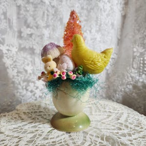 May include: A decorative Easter egg cup with a yellow chick, a small bunny, and a miniature orange tree. The arrangement includes a purple mushroom, speckled eggs, and green decorative accents. The egg cup is cream-colored with a green base.