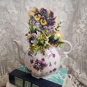 May include: A white porcelain teapot with purple floral patterns, adorned with artificial flowers, Easter eggs, and a small bunny figurine. A decorative floral arrangement with pansies sits atop the teapot, which rests on a stack of books. The teapot is a decorative item.