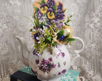 May include: A white porcelain teapot decorated with purple floral patterns, filled with artificial flowers, Easter eggs, and a small bunny figurine. A decorative floral arrangement with a card featuring pansies sits atop the teapot, which rests on a stack of books.