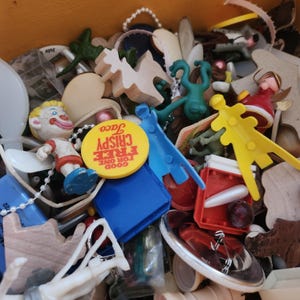 May include: A collection of small toys and trinkets in a wooden box. Items include a clown figurine, a yellow button with text, wooden cutouts, and various plastic shapes in blue, green, and red. The items are scattered and overlapping.