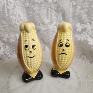 May include: A pair of vintage ceramic salt and pepper shakers shaped like corn on the cob. Each shaker has a smiling or sad face, with black shoes and a yellow collar. The shakers are yellow, brown, and black.