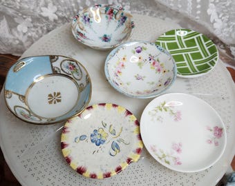 May include: A collection of five small, round ceramic dishes featuring various designs. One dish has a light blue border with gold accents, another has a green and white plaid pattern, and three feature delicate floral motifs in pink, blue, and yellow.